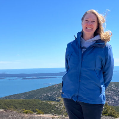 Headshot of Laura Misenheimer, smiling at the camera as she poses near a mountain summit in Acadia on a blue-sky day