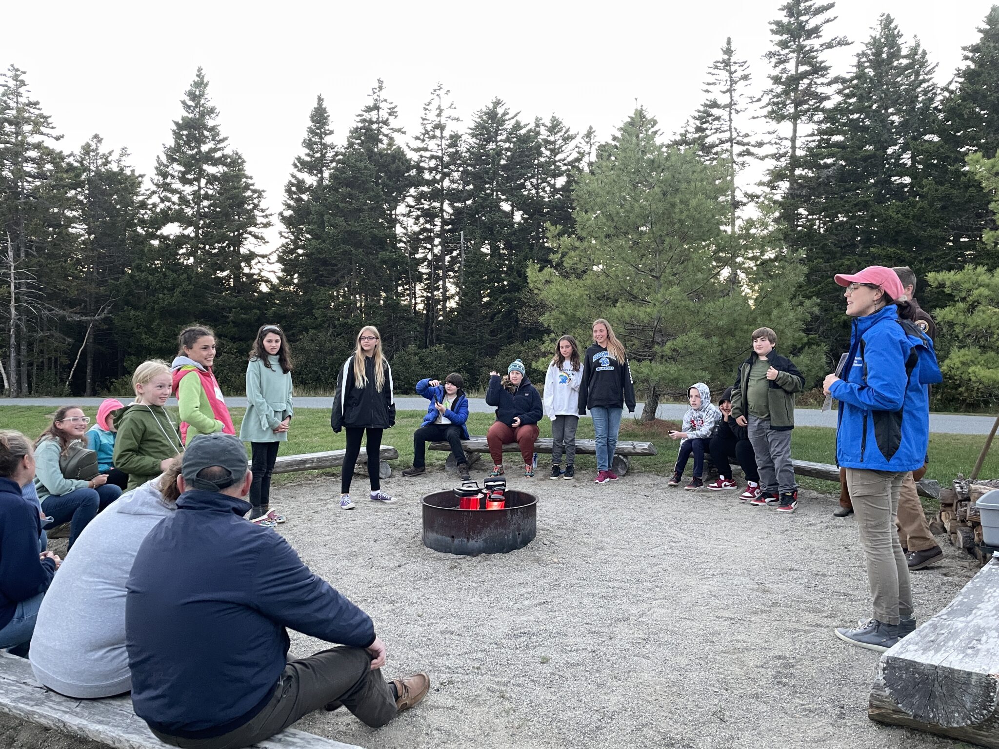 Middle school students, along with their teachers and community chaperones, gather around an outdoor fire pit and listen enthusiastically to an environmental educator