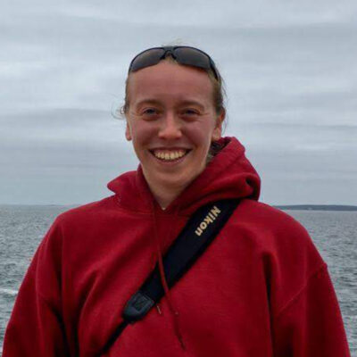 Val Watson wears a red sweatshirt with a camera strap slung over her shoulder and smiles, with the moody ocean behind her