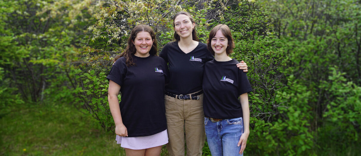 Standing from left to right with arms linked are Abby Schill, Gillian Audier, and Julia Rush, posing for the camera in front of leafing trees and bushes
