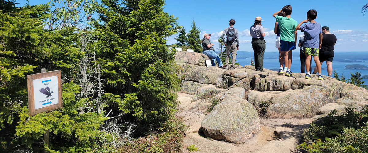 A group of young kids gather with volunteers near the summit of Cadillac Mountain in Acadia National Park to monitor hawk migration under a clear blue sky