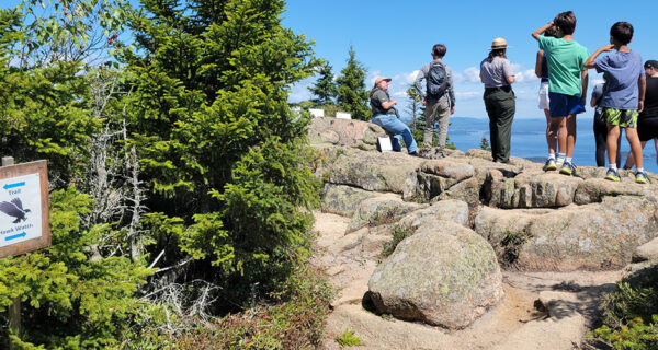 A group of young kids gather with volunteers near the summit of Cadillac Mountain in Acadia National Park to monitor hawk migration under a clear blue sky