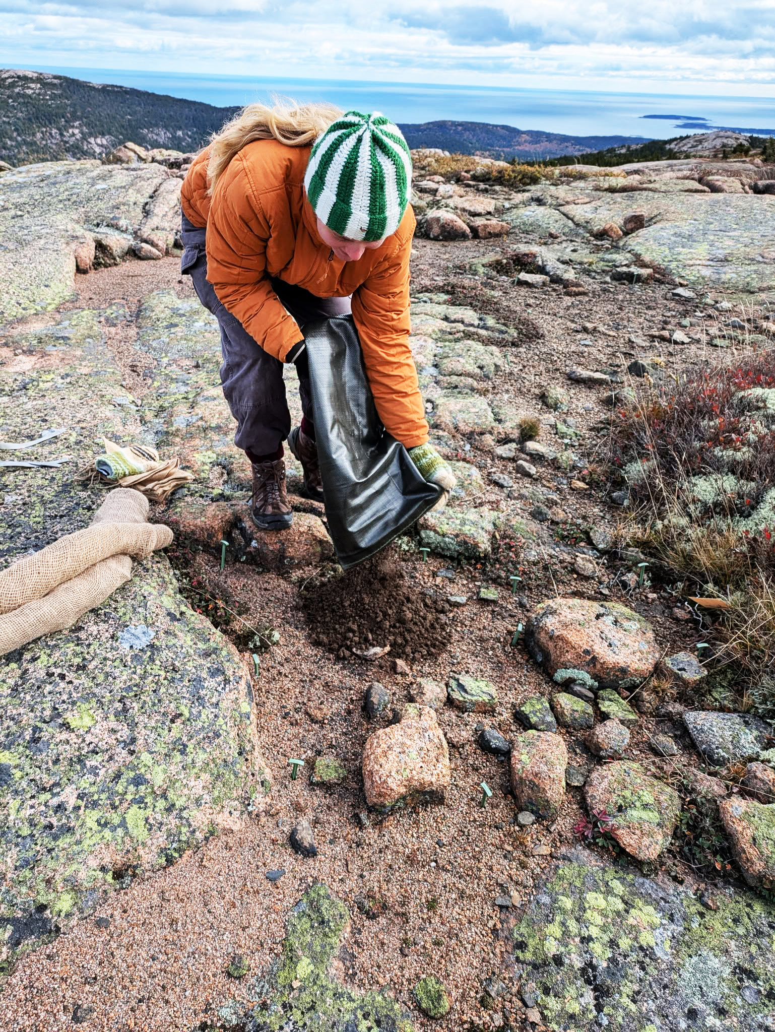 A person empties a bag of soil onto the ground of a mountain summit