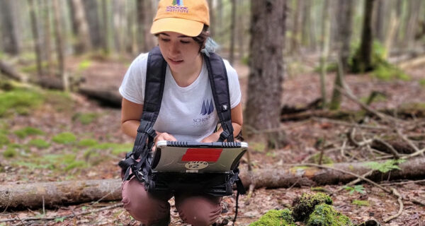 Lucy crouches low to the ground while holding a tablet and observing the forest floor