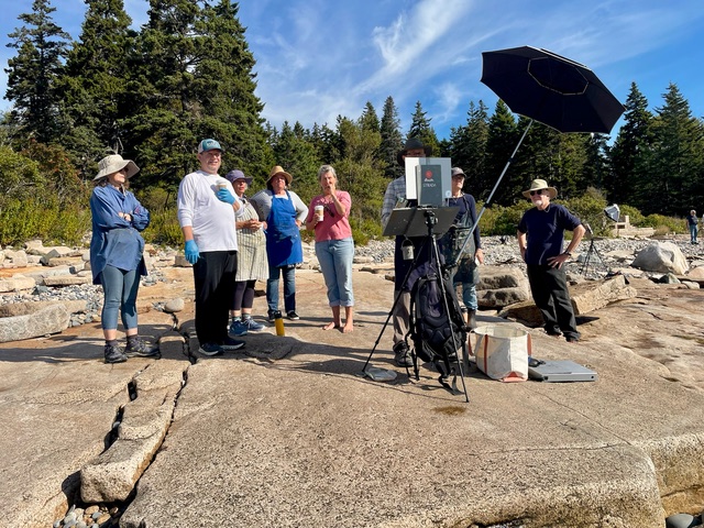A group of painters stand on the rocky coastline while observing a painting on an easel