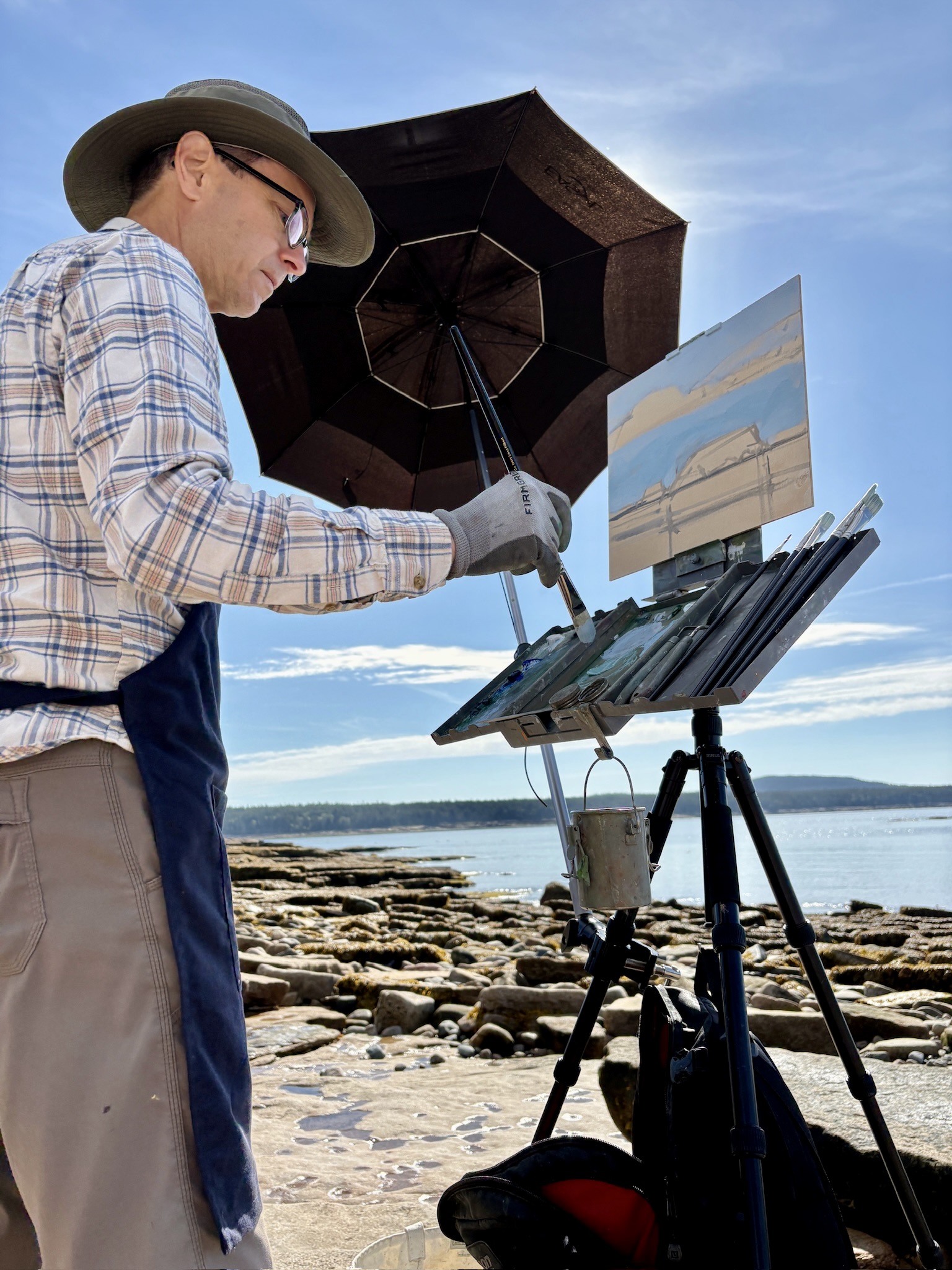 Philip Frey stands at his easel while painting under an umbrella on a bright, sunny day