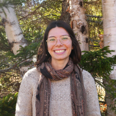 Molly Donlan poses for a photo in front of fir and birch trees, with the sunlight peaking through the branches