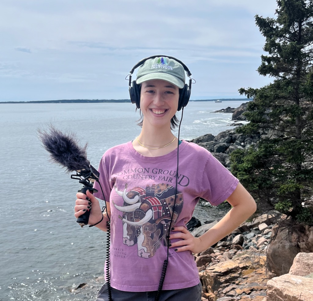Julia Rush holds podcasting equipment while smiling for the camera near the coastline 